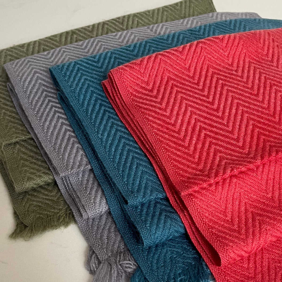 Four folded woven cashmere scarves with a herringbone pattern, stacked diagonally. The visible colors are olive green, light gray, teal blue, and bright coral red.